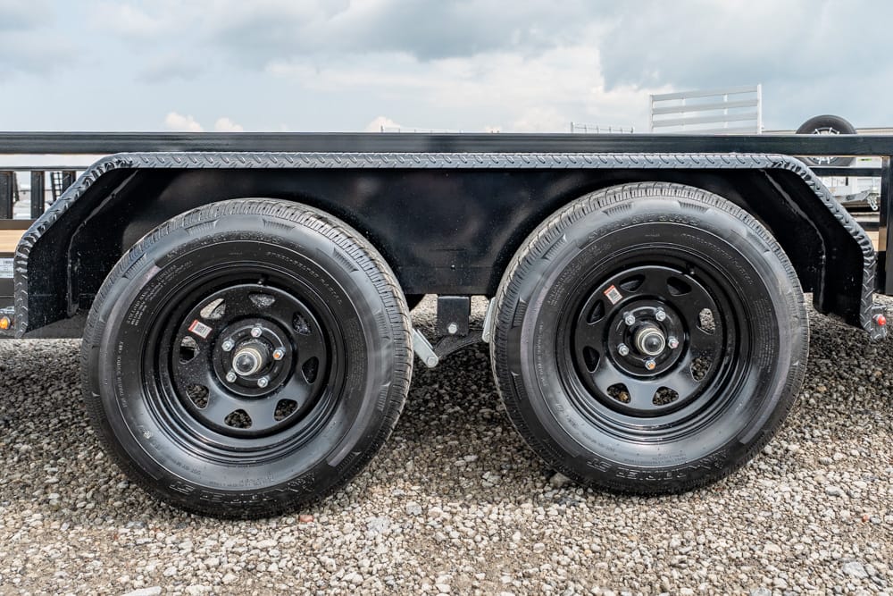 Close-up of dual trailer wheels with black rims on a gravel surface under a black fender and diamond-plate edge.