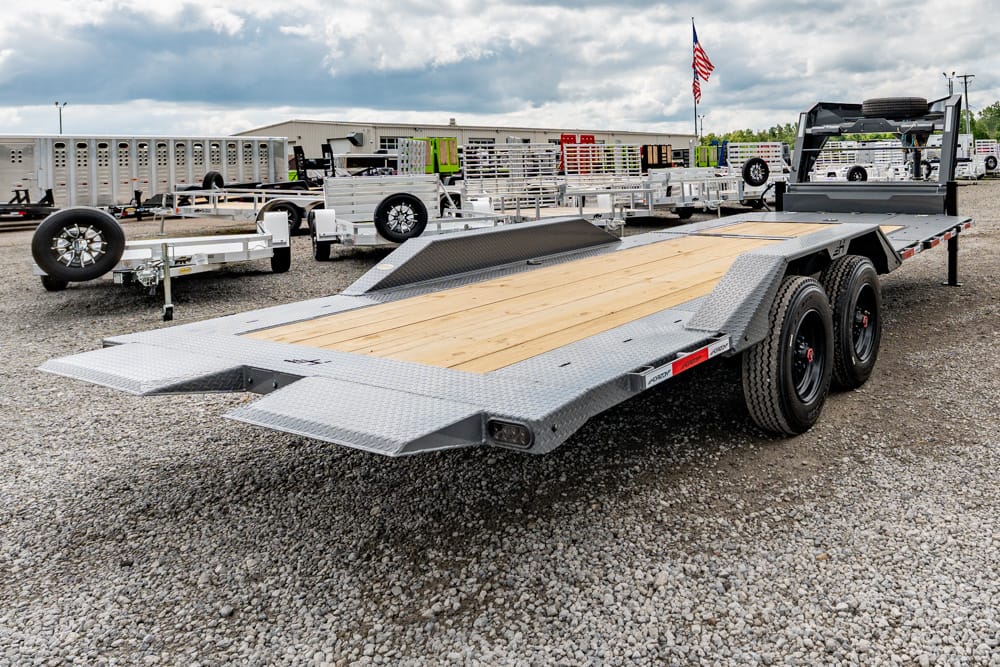 Flatbed car trailer with a wooden deck and diamond-plate steel ramps in a gravel lot among other trailers and equipment.",