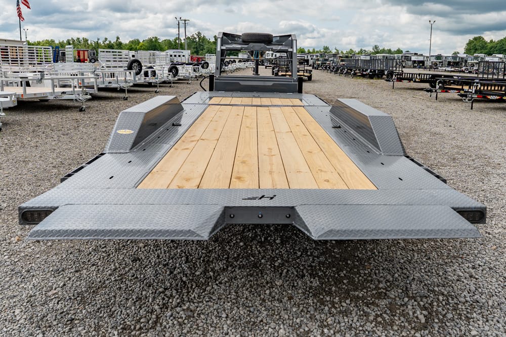 Flatbed tow trailer with a wooden deck and diamond-plate steel ramps in a gravel lot.