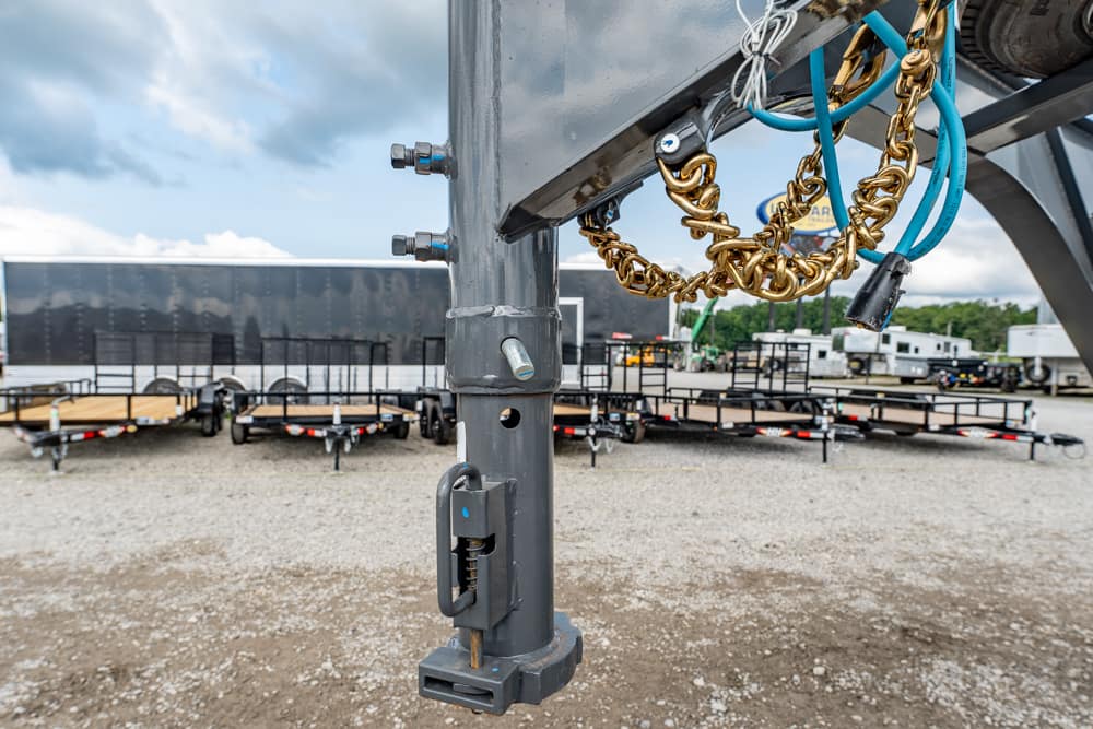 Close-up of a gooseneck trailer hitch with safety chains, blue hoses, and a ground jack in a gravel lot.