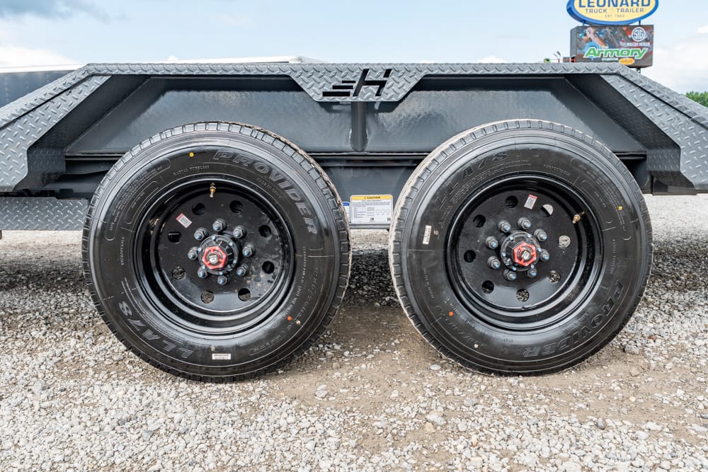 Dual-axle trailer with black steel wheels and heavy tires on a gravel surface, with diamond-plate fenders above the wheels.