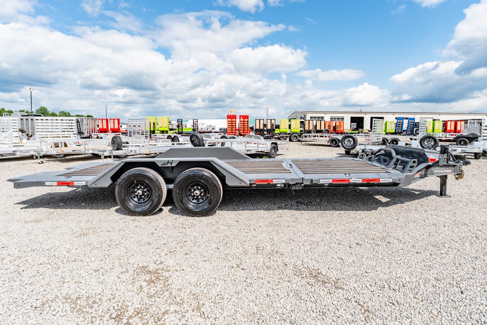 Gray dual-axle car hauler trailer on a gravel lot with other colorful trailers in the background
