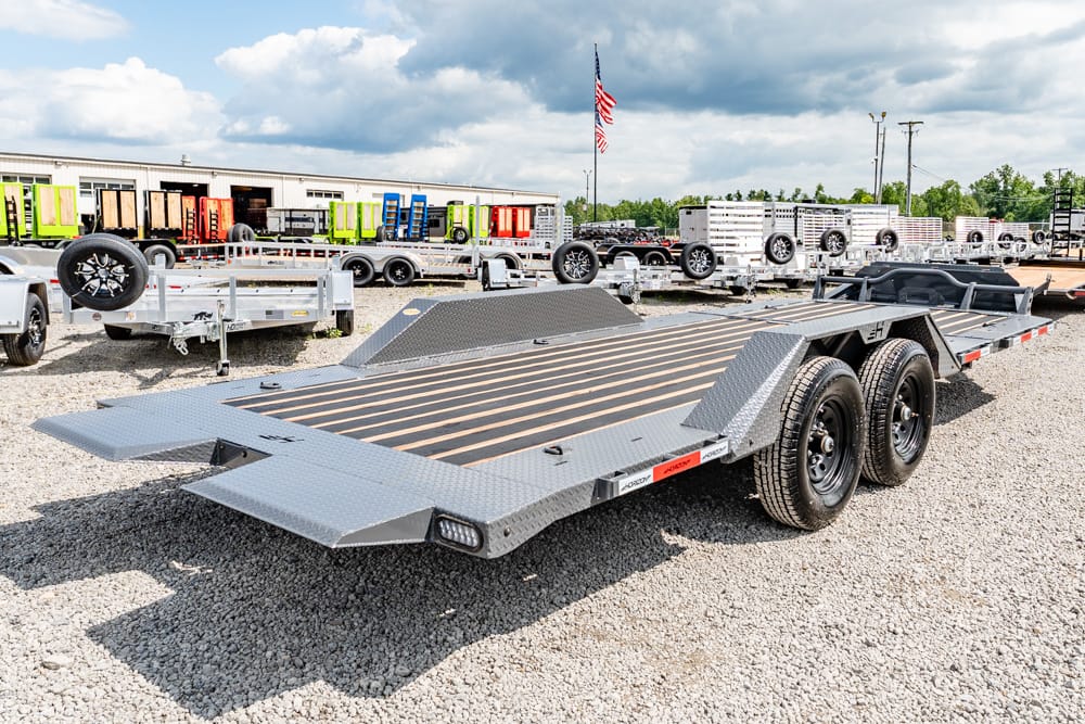 Flatbed car hauler trailer with dual axles and wooden loading deck parked on gravel at a trailer lot.
