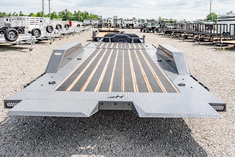 Wide view of a metal flatbed car hauler trailer with wooden deck planks, diamond-plate ramps at the rear, parked on a gravel lot with other trailers in the background