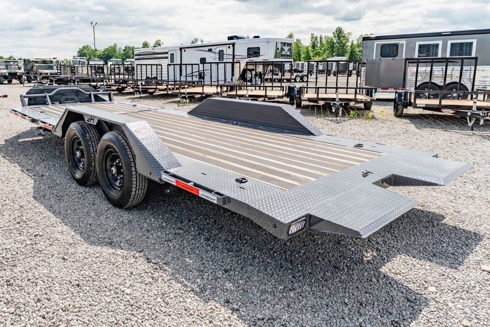 Double-axle flatbed car hauler trailer with folding ramps on a gravel lot, with RVs and trailers in the background.