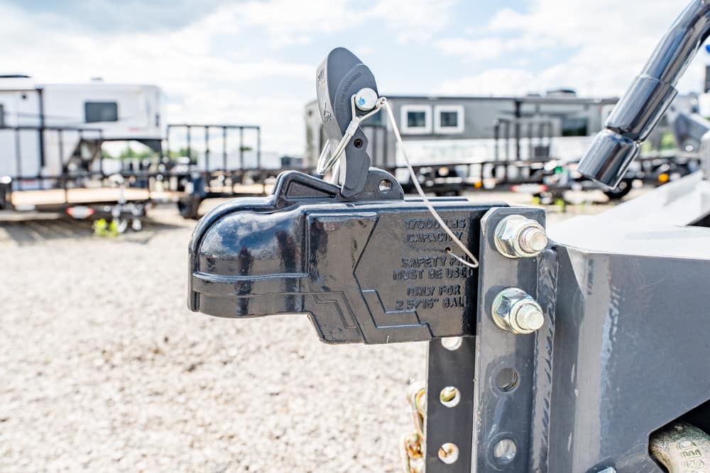 Close-up of a black trailer coupler and hitch latch on a metal trailer frame, with a safety pin and chain, outdoors on gravel with RVs in the background