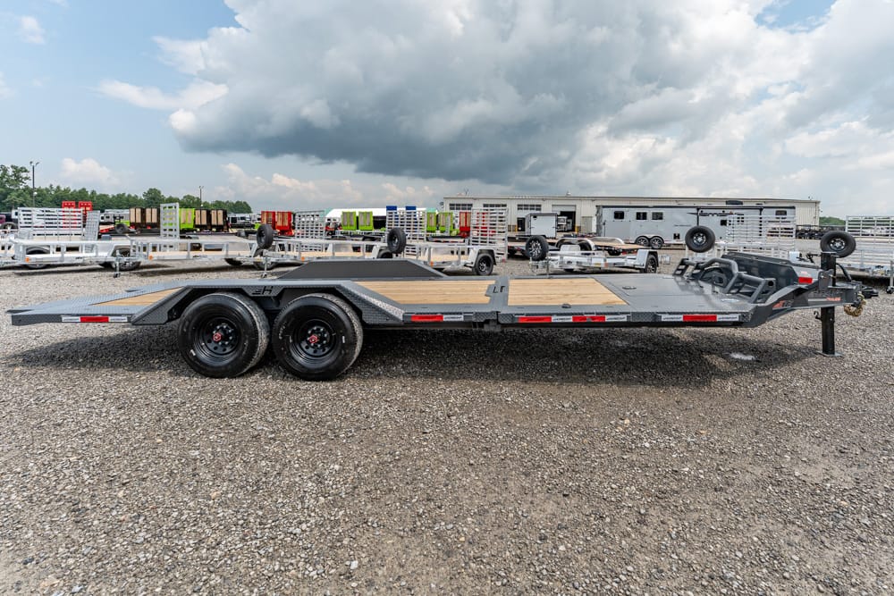 Flatbed trailer with dual axles and a wooden deck parked on a gravel lot, with other trailers and a cloudy sky in the background.