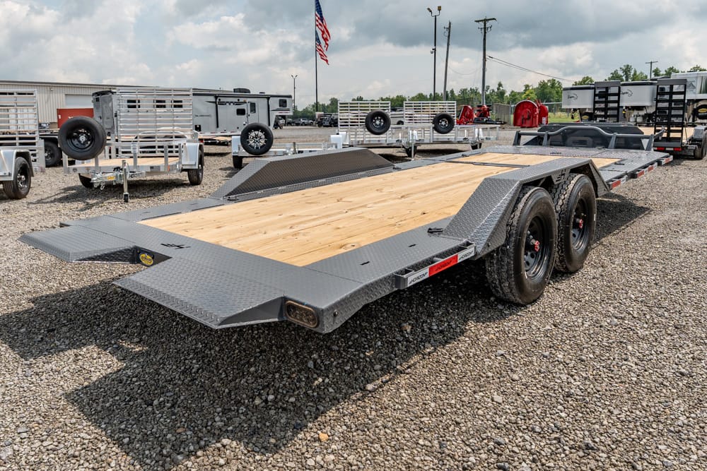 Flatbed car trailer with a wooden deck and metal ramp sections sits on a gravel lot, ready for transport use.