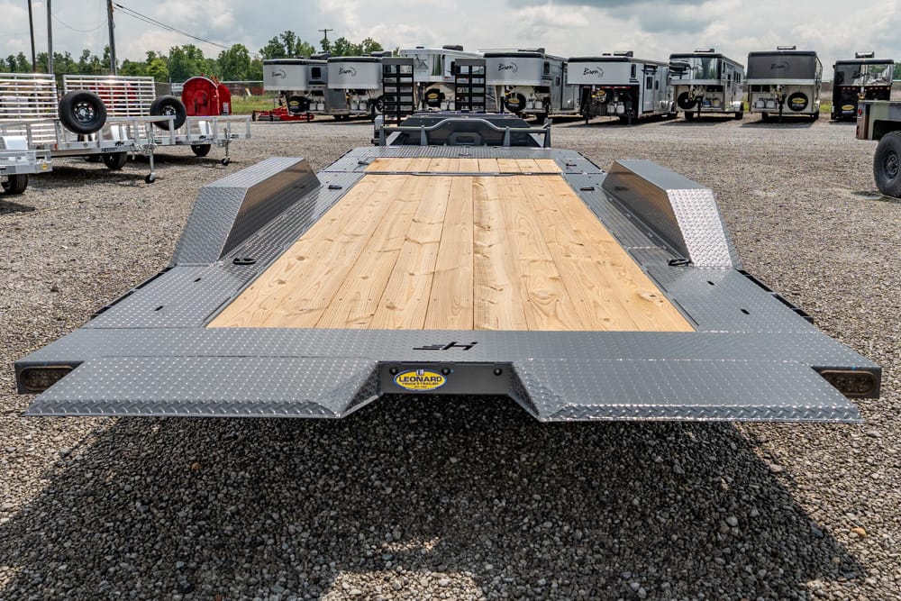 Flatbed trailer with a wooden deck and diamond-plate steel ramps in a gravel lot, other trailers visible in the background, likely at a trailer yard.