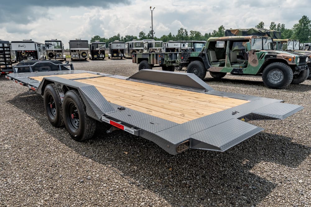 Heavy-duty flatbed trailer with wooden deck and diamond-plate ramps in a gravel lot, backed by trucks and military jeeps.