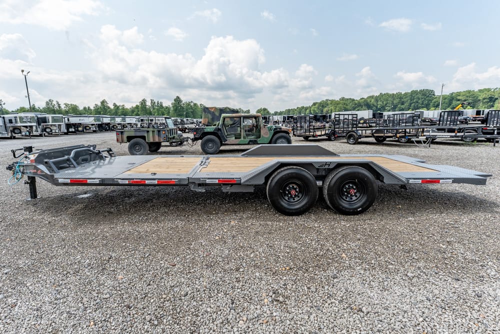 Dual-axle steel car hauler trailer with a wooden deck on a gravel lot, other trailers in the background.