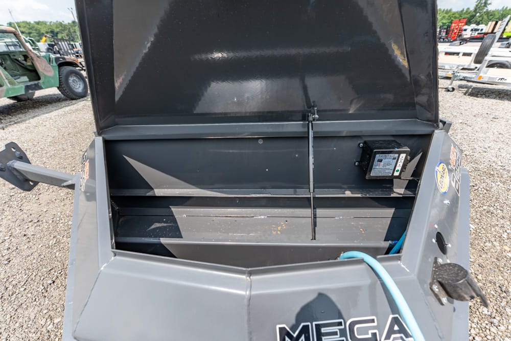 Open metal storage bin on a gray industrial machine labeled MEGA; blue hose rests across the front and a small control box is mounted on the right side in a gravel lot.