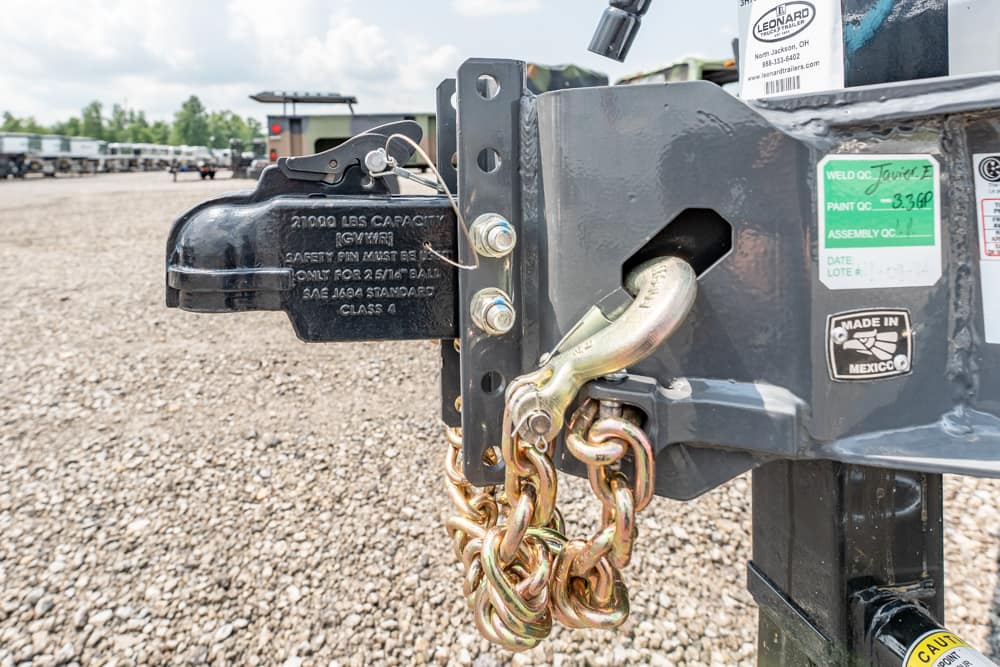 Close-up of a trailer hitch with a locking mechanism and tangled chains attached to a gray metal frame on a gravel lot