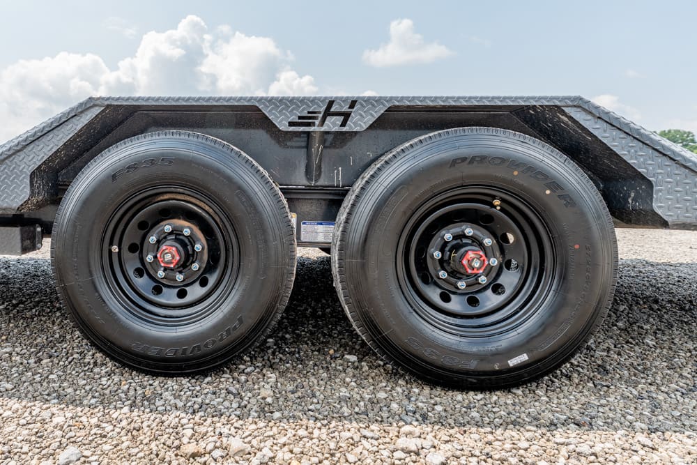 Dual-axle trailer with two black steel wheels on a gravel lot under a metal fender.