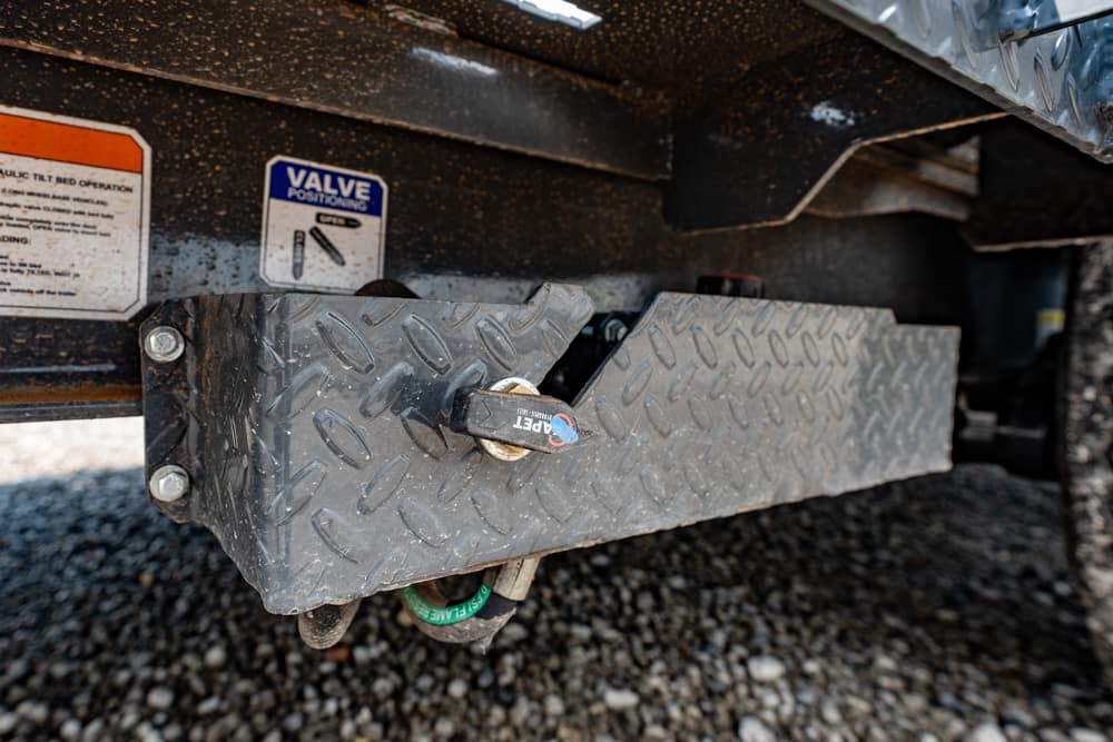 Close-up of a diamond-plate toolbox under a trailer, showing a latch and a green wheel stud on gravel behind it.