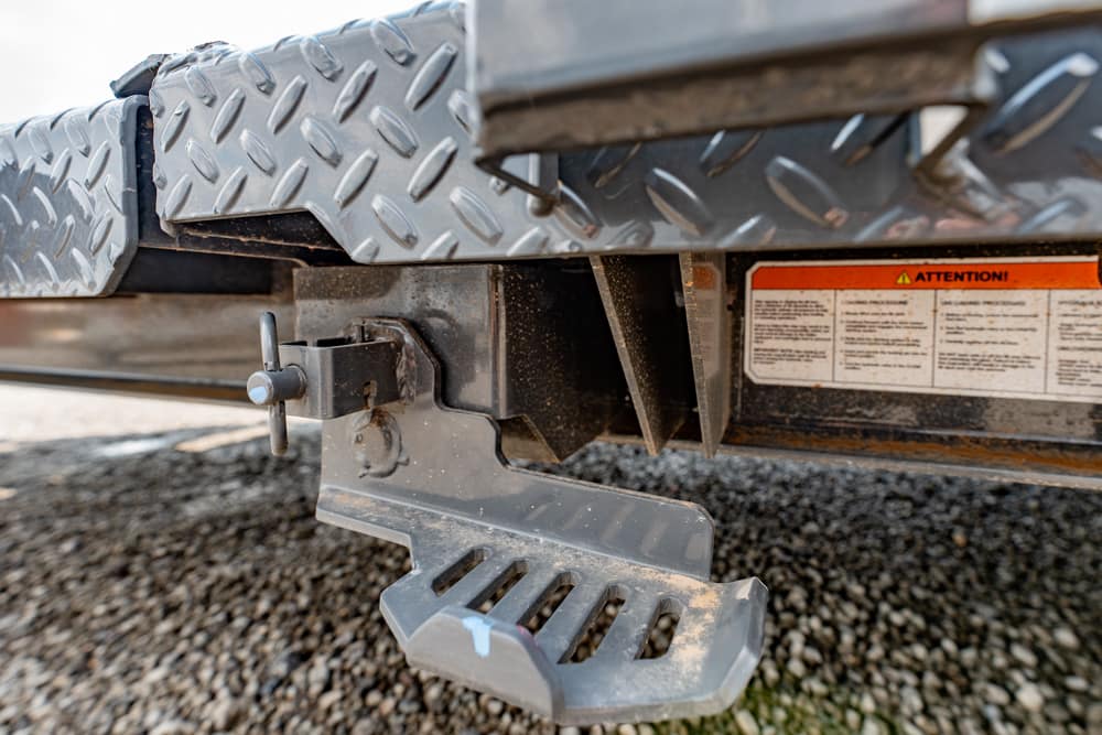 Close-up of a trailer hitch and built-in step with diamond-plate surface on a gravel surface.