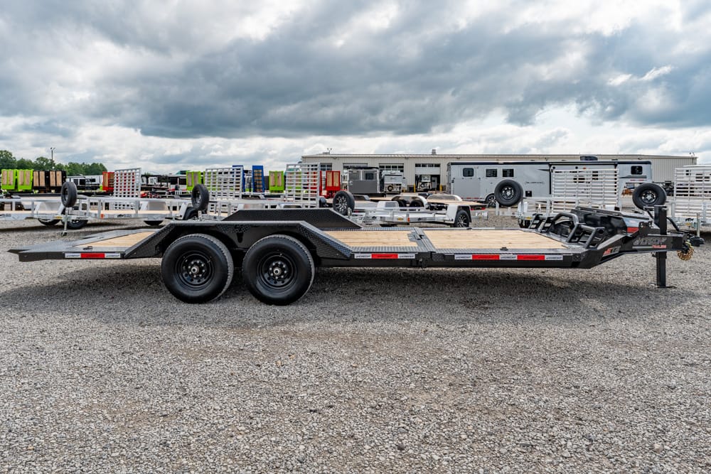 Black tandem-axle car trailer with a wooden deck sits on a gravel lot, surrounded by other trailers and a warehouse in the background.