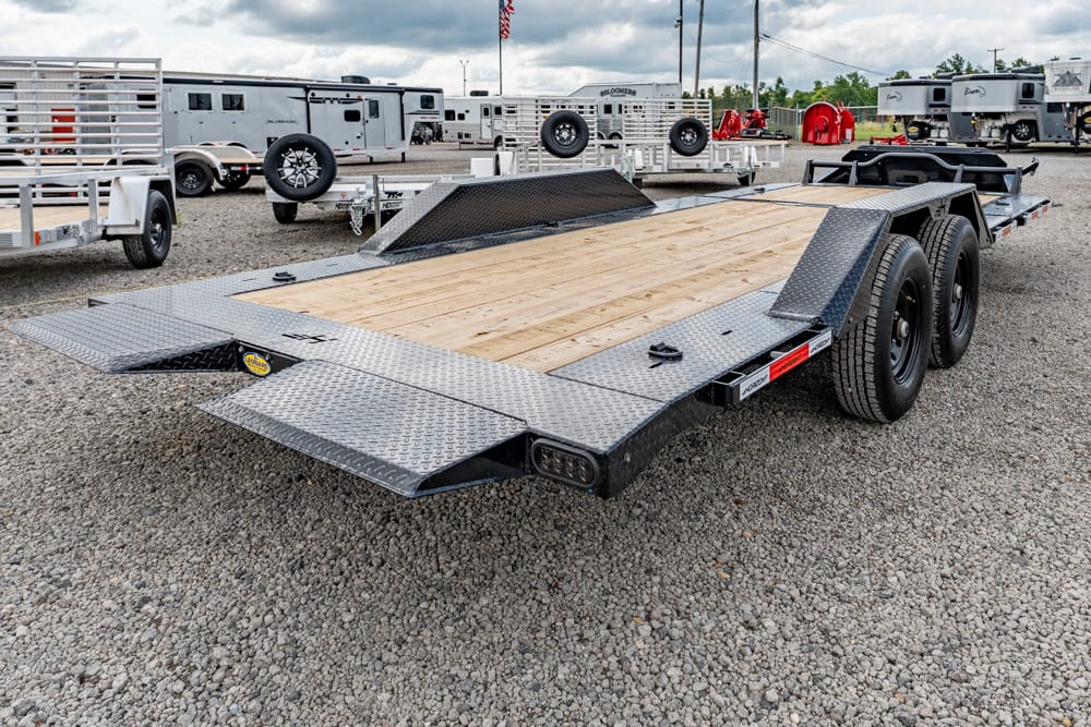 Two‑axle steel car hauler with wooden deck and diamond‑plate ramps on a gravel lot, surrounded by other trailers and equipment