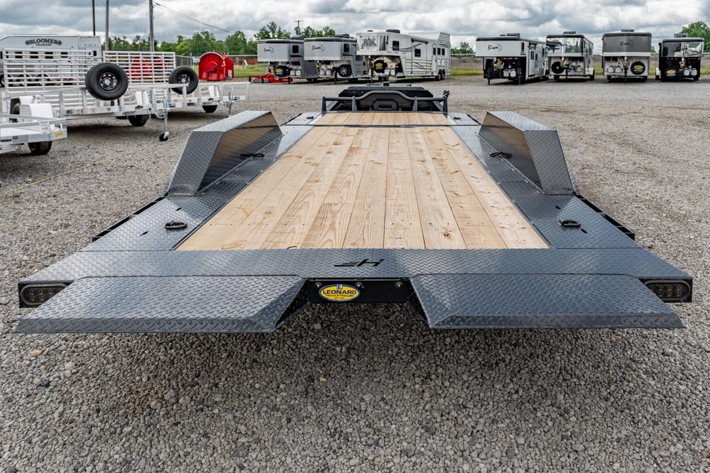 Flatbed car-hauler trailer with a wooden deck and diamond-plate steel ramps, parked on a gravel lot with travel trailers in the background.