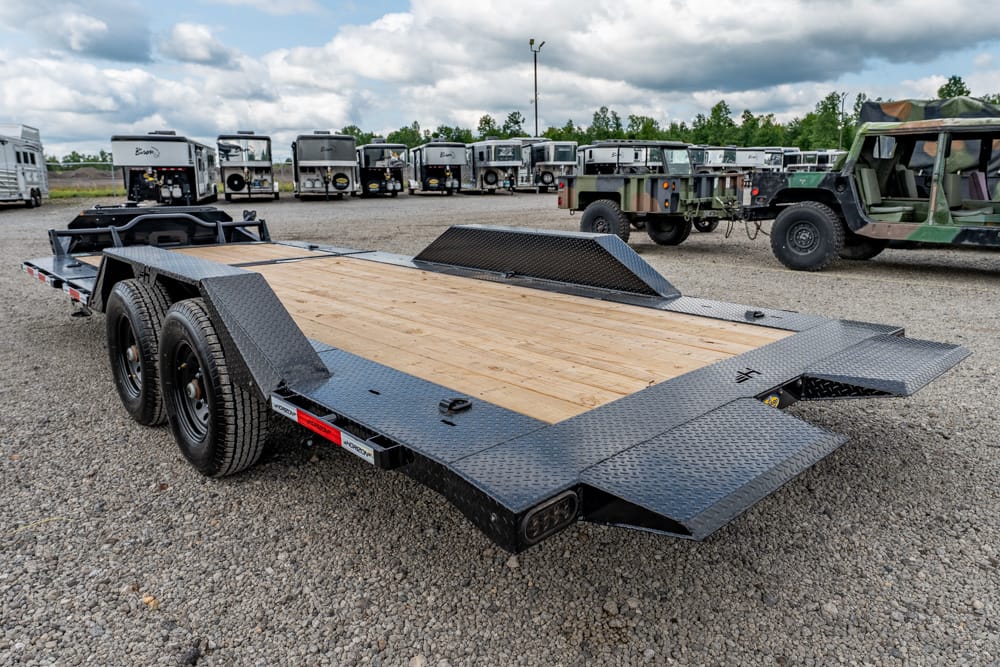 Heavy-duty car hauler trailer with a wooden deck and diamond-plate ramps in a gravel lot among other trailers and military vehicles.