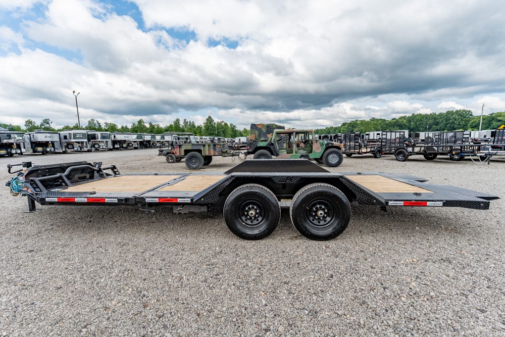 Flatbed car trailer with a wooden deck and dual axles on a gravel lot, with military trucks in the background.