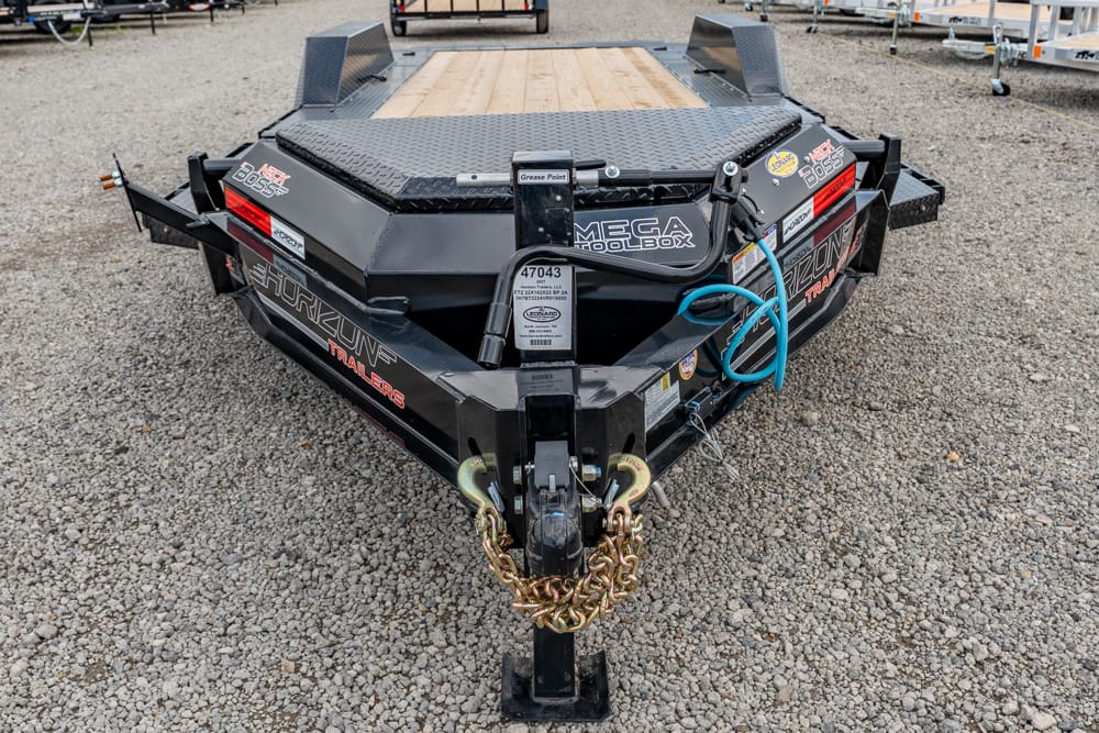 Front view of a black heavy-duty trailer hitch on a gravel lot, with safety chains, coiled blue hose, and a wood deck surface visible on top of the trailer.