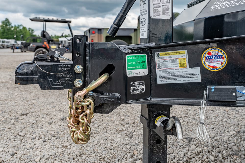 Close-up of a trailer hitch with a thick safety chain, latch hardware, and label stickers on a black metal frame.