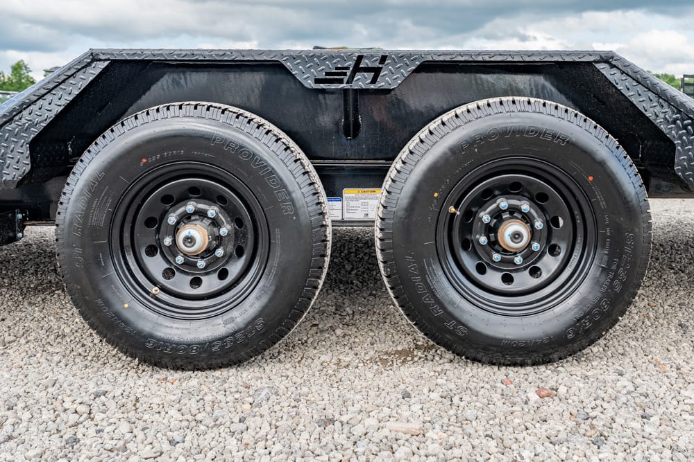Dual trailer wheels with black rims and lug nuts under a diamond-plate fender on a gravel surface, showing a tandem-axle setup.