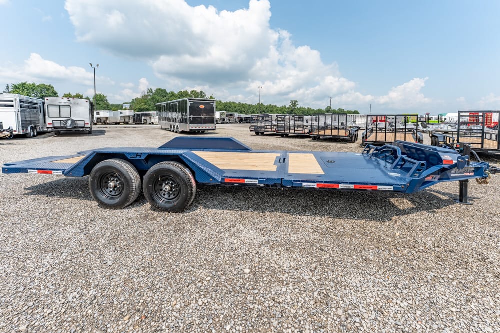 Blue dual-axle car hauler trailer on a gravel lot, with wooden deck and fold-down ramps at the rear edge of the trailer's bed