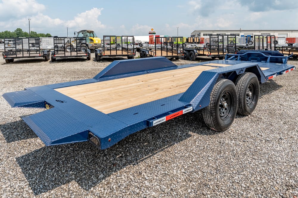 Blue two-axle flatbed trailer with a wooden deck parked on a gravel lot; folded ramp extensions at the ends and other trailers visible in the background.