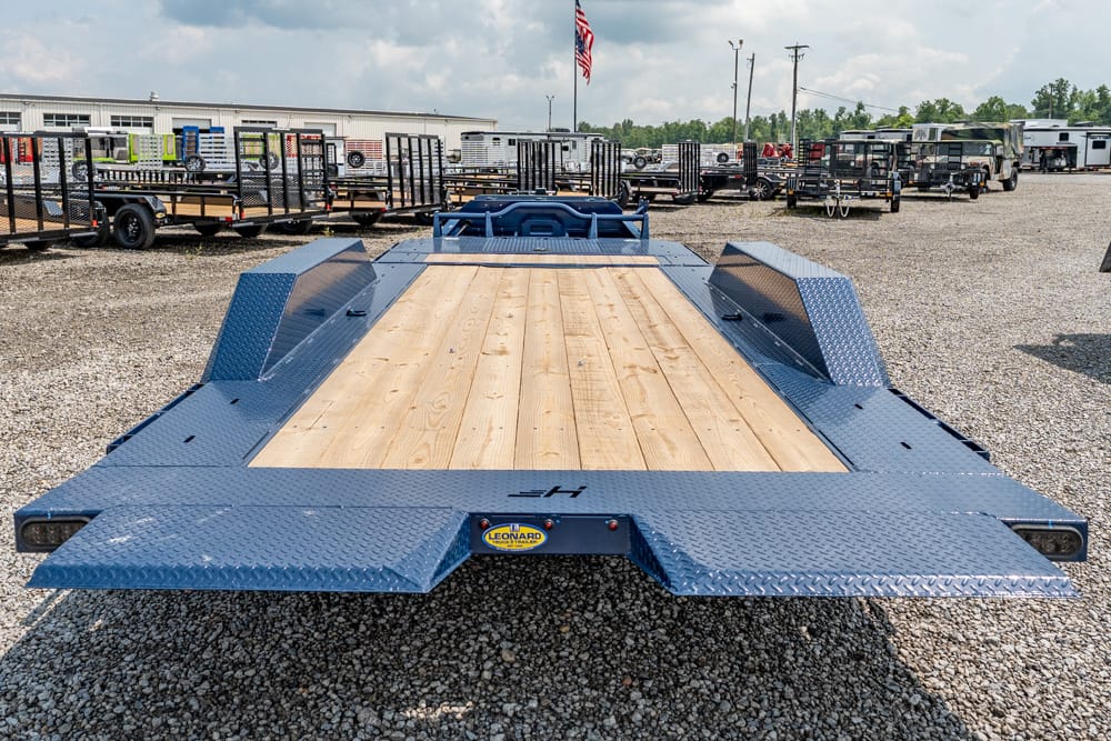 Blue heavy-duty flatbed trailer with wooden deck, front view, diamond-plate fenders, Leonard logo on hitch, parked on gravel lot with other trailers in background