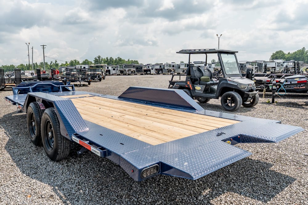 Blue heavy-duty flatbed trailer with a wooden deck, parked on a gravel lot with other trailers in the background and a utility cart nearby.
