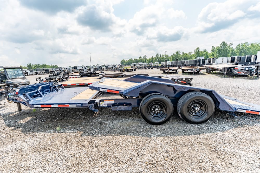 Blue flatbed car hauler trailer with dual rear wheels in a gravel lot among many trailers in the background.