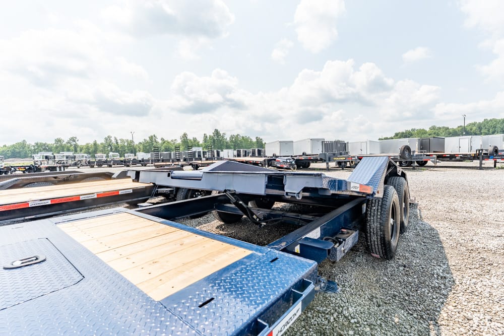 Blue metal flatbed trailer with wooden decking, parked in a gravel trailer yard with many trailers in the background.