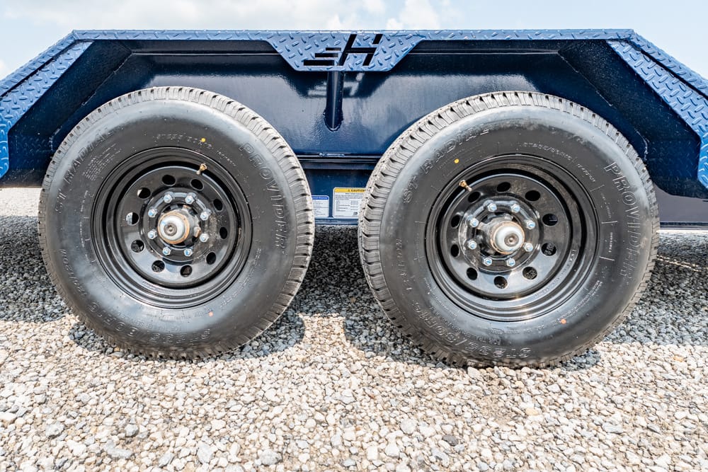 Pair of black trailer wheels with lug nuts on a blue utility trailer, resting on gravel under the fender.