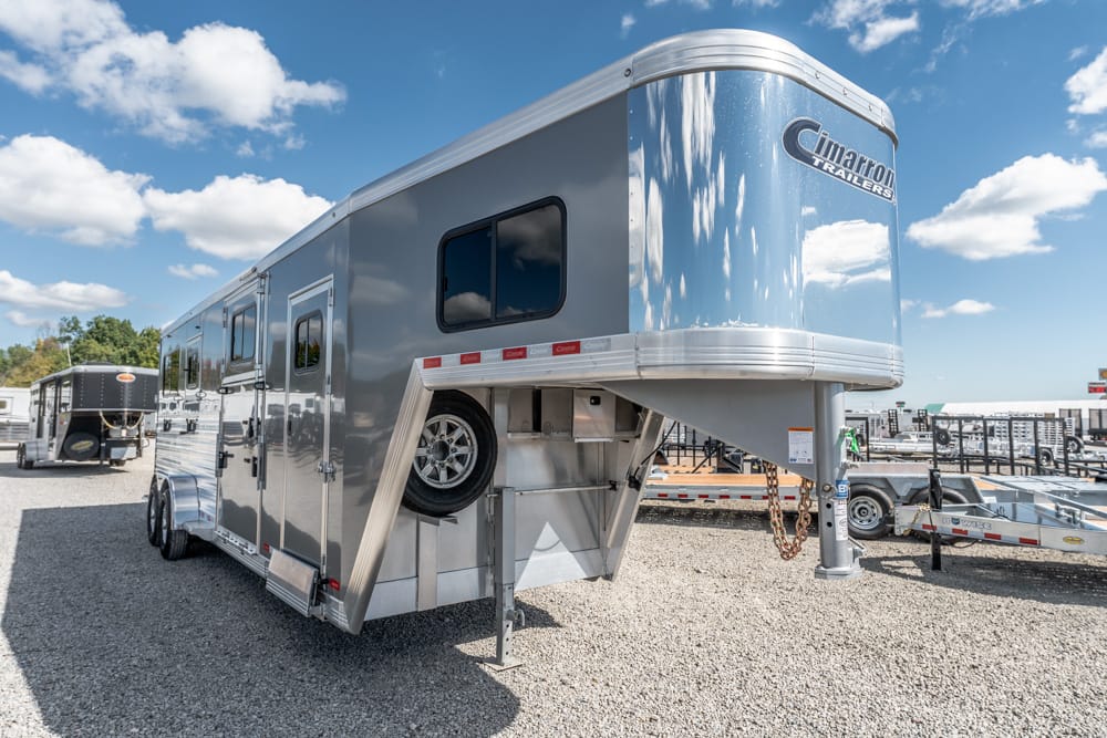Gooseneck adventure trailer in a gravel lot, side view showing a Cimarron trailer with a spare tire mounted on the side and chrome trim.