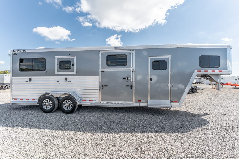 Silver gooseneck horse trailer with two axles and multiple entry doors parked on a gravel lot under a blue sky with scattered clouds, side view. The trailer has several windows and metal siding.