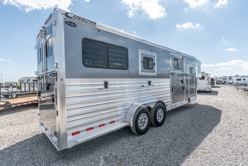 Silver aluminum, dual-axle horse trailer parked on a gravel lot with side windows and doors visible in bright daylight.