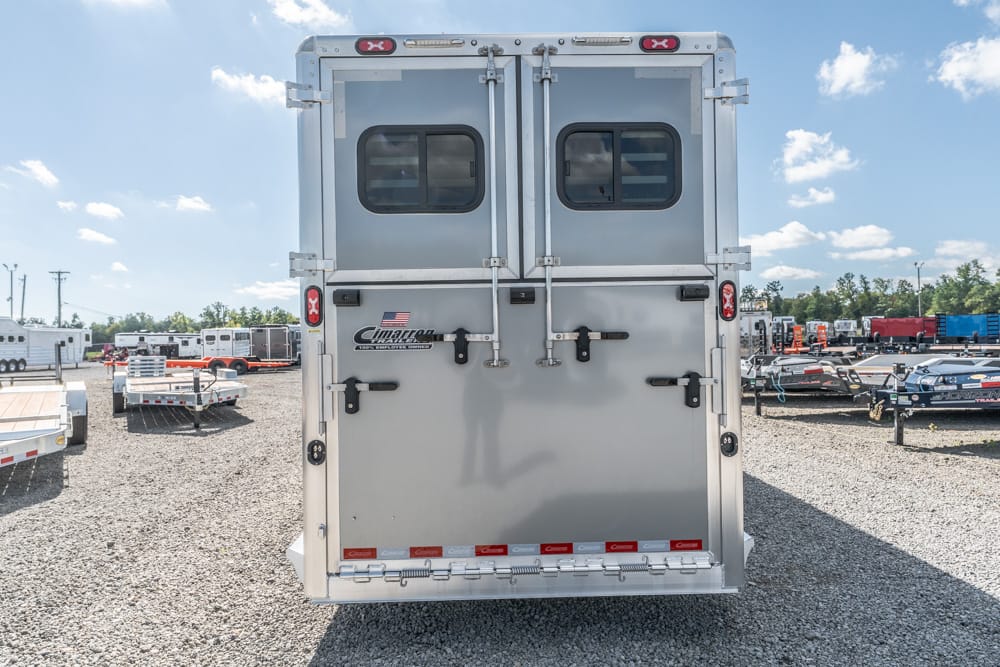 Rear view of a silver enclosed cargo trailer with double doors and latches in a gravel lot, other trailers and vehicles in the background.
