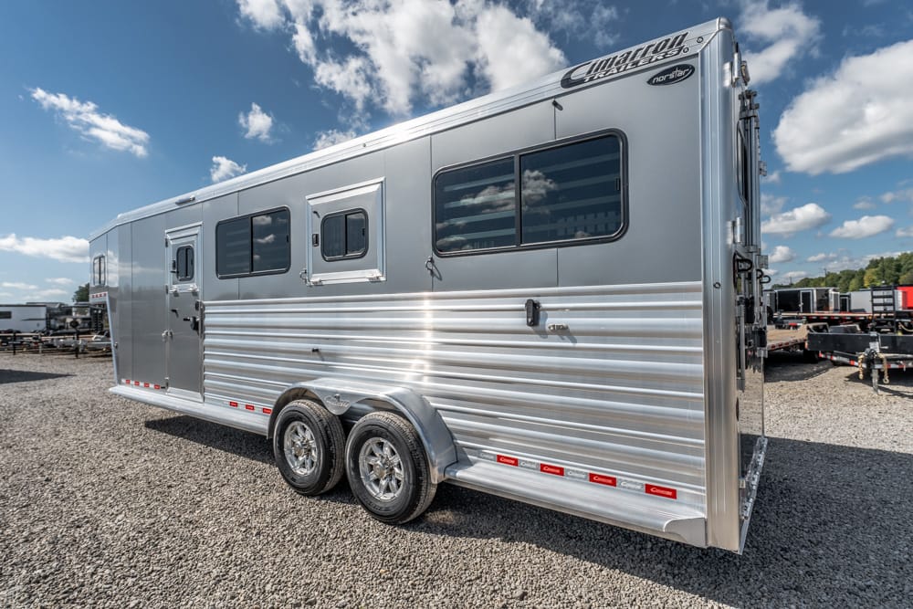 Silver double-axle travel trailer parked on a gravel lot, showing side windows and ribbed aluminum siding under a blue sky.