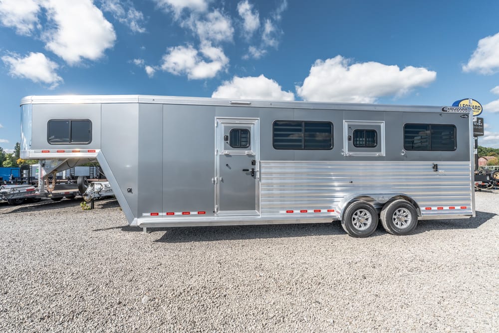 Silver aluminum gooseneck horse trailer with two axles, windows, and front door parked on a gravel lot under a blue sky.