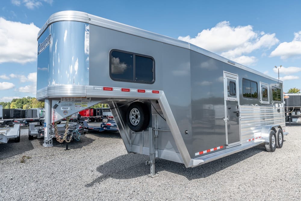 Gray gooseneck horse trailer with raised front hitch and spare tire, parked on a gravel lot under a blue sky.