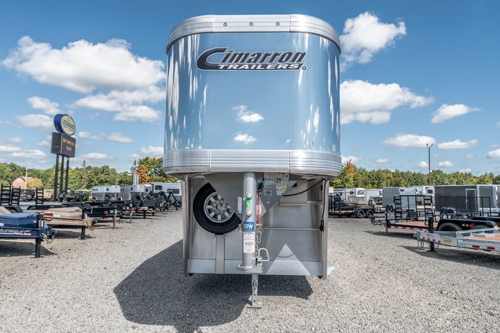 Shiny aluminum Cimarron gooseneck trailer in a gravel trailer lot under a blue sky with clouds, surrounded by other trailers.