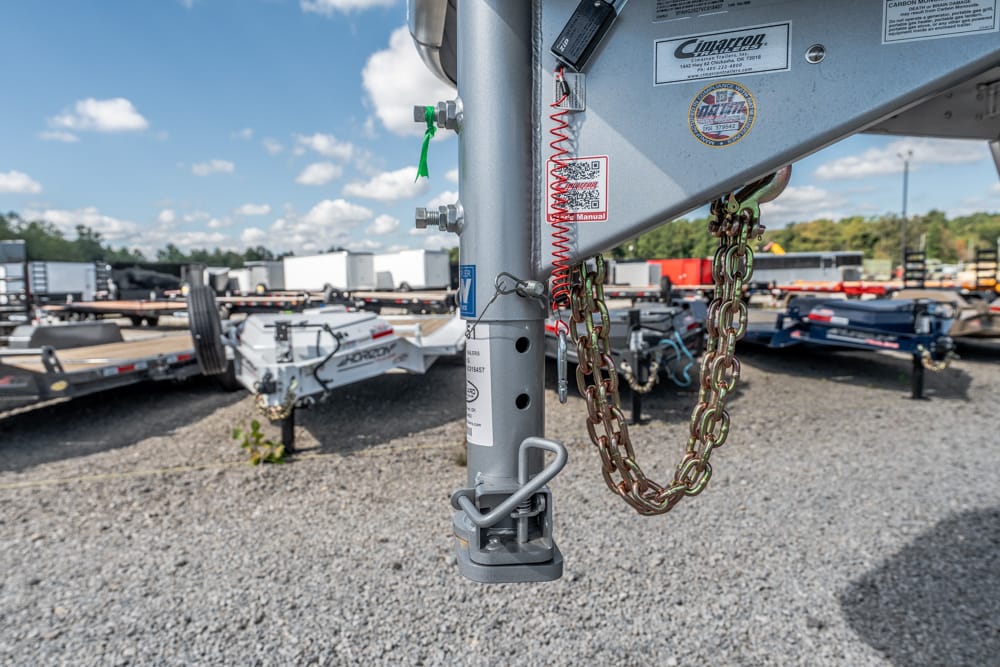 Close-up of a trailer hitch with safety chains and labels, in a gravel lot with trailers in the background.