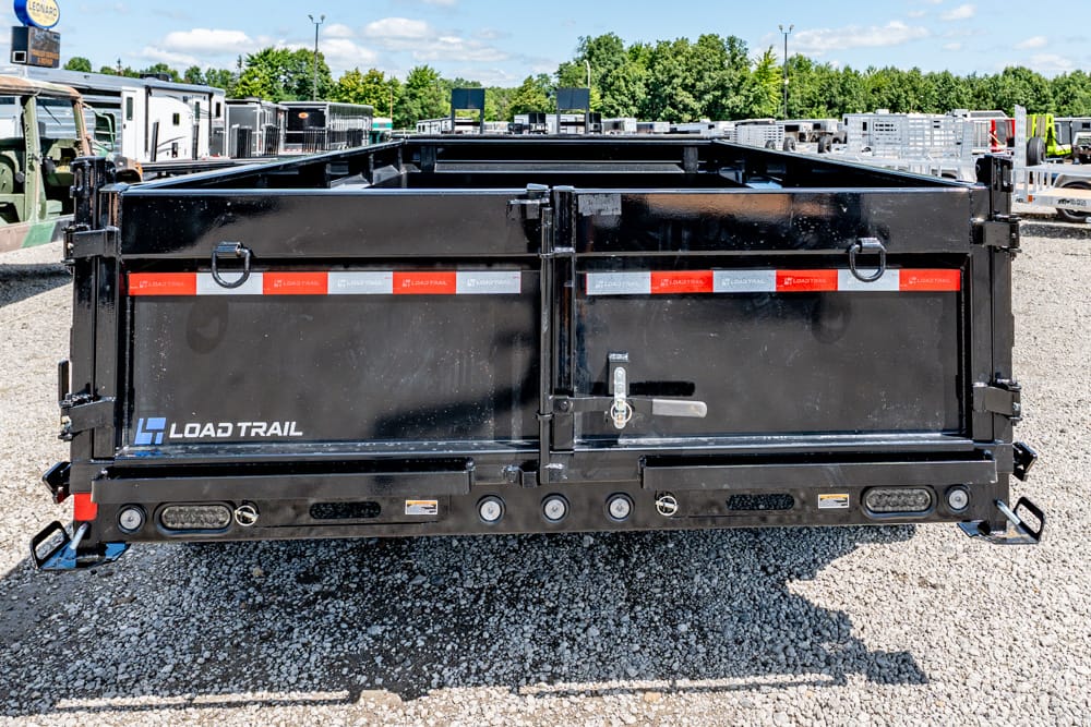 Rear view of a black Load Trail dump trailer in a gravel lot, showing latches and taillights on the back doors
