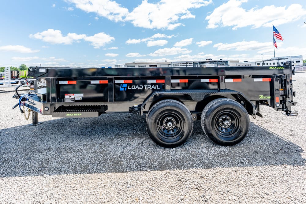 Black dual-axle dump trailer with Load Trail branding on a gravel lot, American flag in background, clear blue sky.