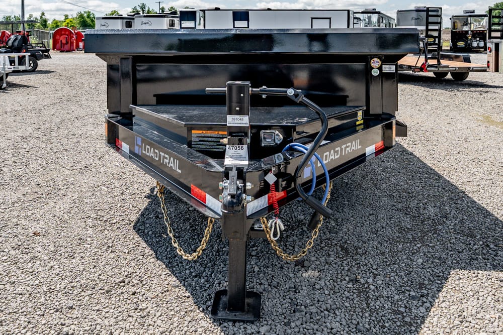Rear view of a black Load Trail utility trailer on a gravel lot, showing the hitch, safety chains, and coiled hoses in front.