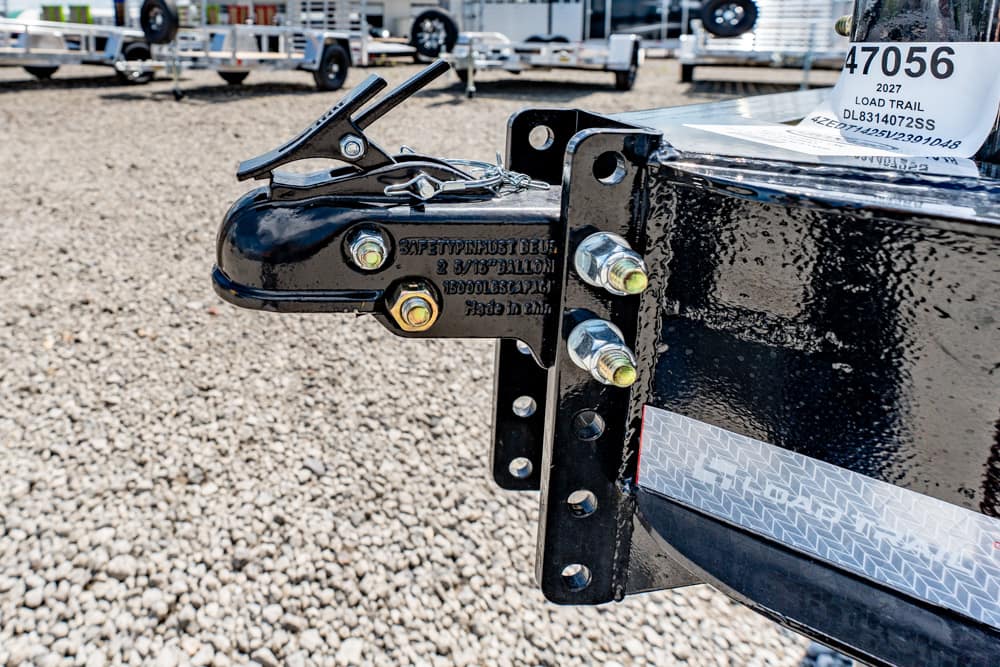 Close-up of a black trailer hitch with safety chains and bolts on a gravel lot, with trailers in the background outdoors in a storage yard.