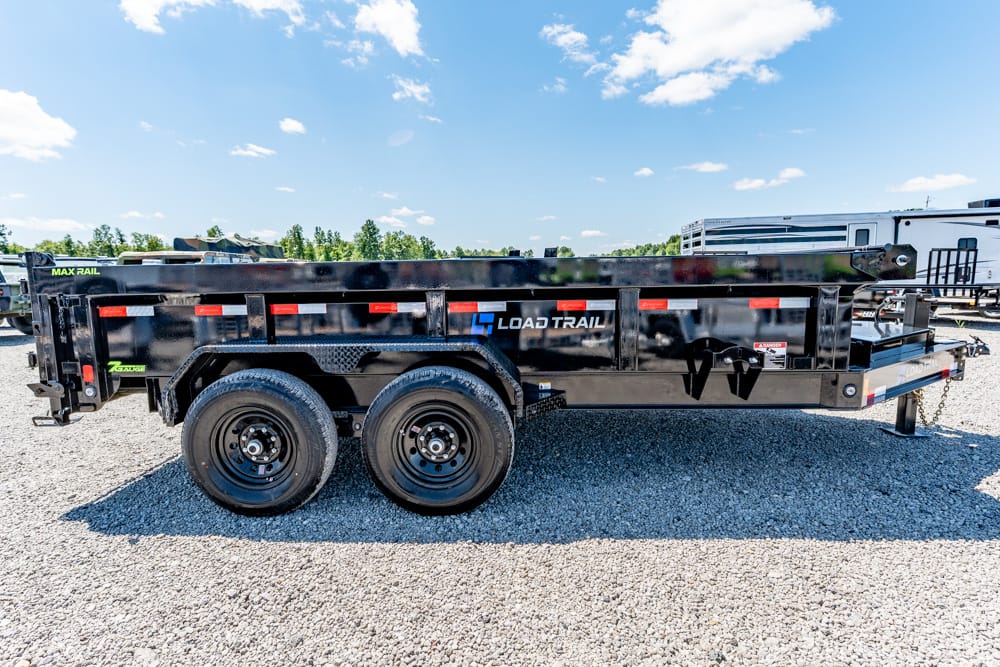 Black Load Trail dump/utility trailer with two axles parked on a gravel lot under a blue sky; red reflective stripes along the sides and a front hitch visible.