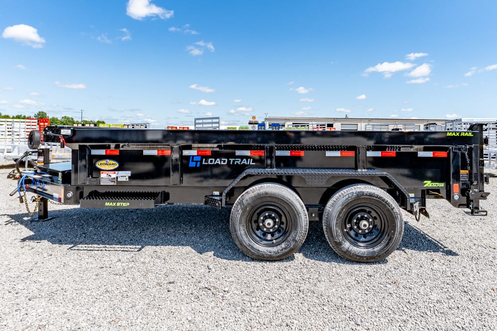 Black Load Trail tandem-axle dump trailer on a gravel lot under a blue sky, side view showing branding and safety decals